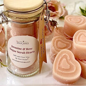 May include: A glass jar with a wooden lid and metal clasp contains pink sugar scrub. Heart-shaped sugar scrub soaps are stacked nearby. The jar has a label that reads "Jasmine & Rose Sugar Scrub Hearts". A handmade tag hangs from the jar.