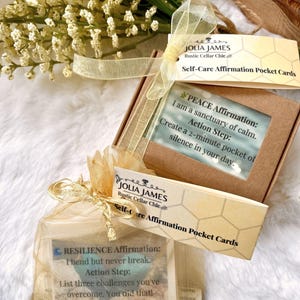 Può includere: Una scatola di cartone marrone e un sacchetto in organza dorata con coulisse contenenti carte di affermazione. L'etichetta della scatola riporta Jolia James Rustic Cellar Chic Self-Care Affirmation Pocket Cards e Peace Affirmation. Il sacchetto riporta Resilience Affirmation.