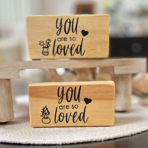 May include: Two wooden blocks with the phrase "you are so loved" in black script. One block has a floral vase graphic, the other has a potted plant graphic. Both blocks have a black heart symbol.