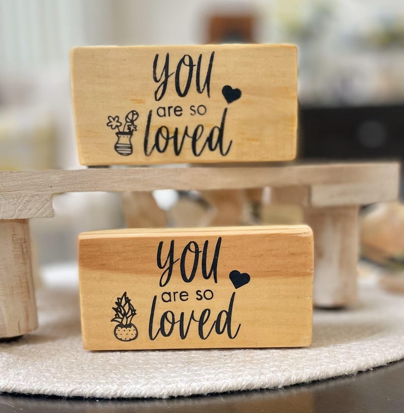 May include: Two wooden blocks with the phrase "you are so loved" in black script. One block has a floral vase graphic, the other has a potted plant graphic. Both blocks have a black heart symbol.