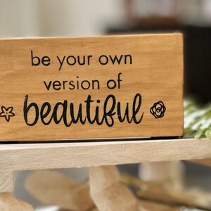 May include: A rectangular wooden block with the phrase "be your own version of beautiful" in black script. A star and a rose graphic are on either side of the word "beautiful". The block is light brown and sits on a light-colored wooden surface.