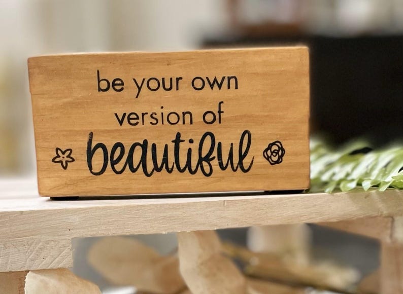 May include: A rectangular wooden block with the phrase "be your own version of beautiful" in black script. A star and a rose graphic are on either side of the word "beautiful". The block is light brown and sits on a light-colored wooden surface.