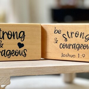 May include: Two wooden blocks with the words "be strong & courageous" in black script. One block has a cactus and heart graphic, the other has a floral design and "Joshua 1:9". The blocks are on a light wood stand.
