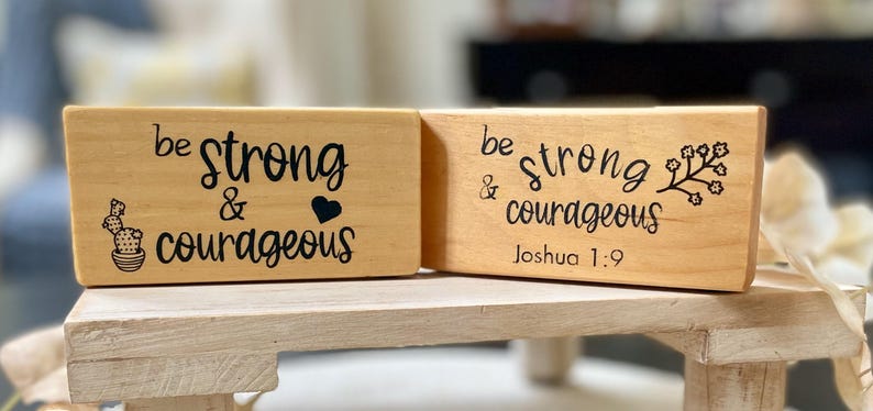 May include: Two wooden blocks with the words "be strong & courageous" in black script. One block has a cactus and heart graphic, the other has a floral design and "Joshua 1:9". The blocks are on a light wood stand.