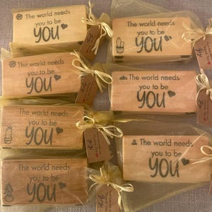 May include: Several small, rectangular wooden blocks with the text "The world needs you to be YOU" are displayed. Each block is wrapped in sheer fabric and tied with a ribbon and a small tag. The blocks have different small illustrations.