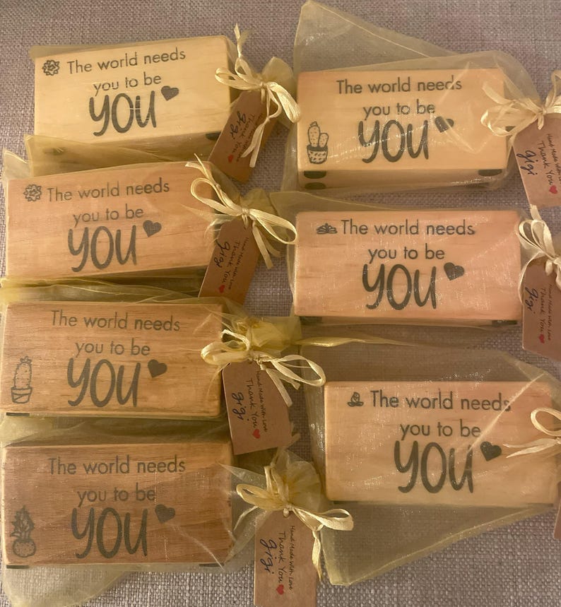 May include: Several small, rectangular wooden blocks with the text "The world needs you to be YOU" are displayed. Each block is wrapped in sheer fabric and tied with a ribbon and a small tag. The blocks have different small illustrations.