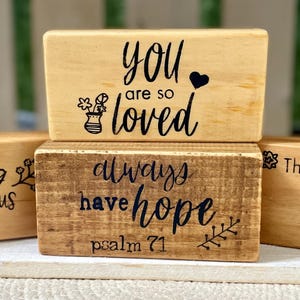 May include: Four wooden blocks with inspirational quotes in black script. The blocks are light brown and dark brown, with phrases like "You are so loved" and "Be strong & courageous".