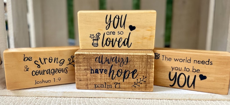 May include: Four wooden blocks with inspirational quotes in black script. The blocks are light brown and dark brown, with phrases like "You are so loved" and "Be strong & courageous".