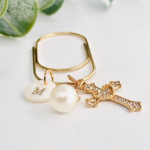 May include: Gold-toned keychain with a cross charm, a pearl, and a circular charm with the letter "M". The cross is encrusted with small, clear crystals. The keychain is set against a white background with green foliage.
