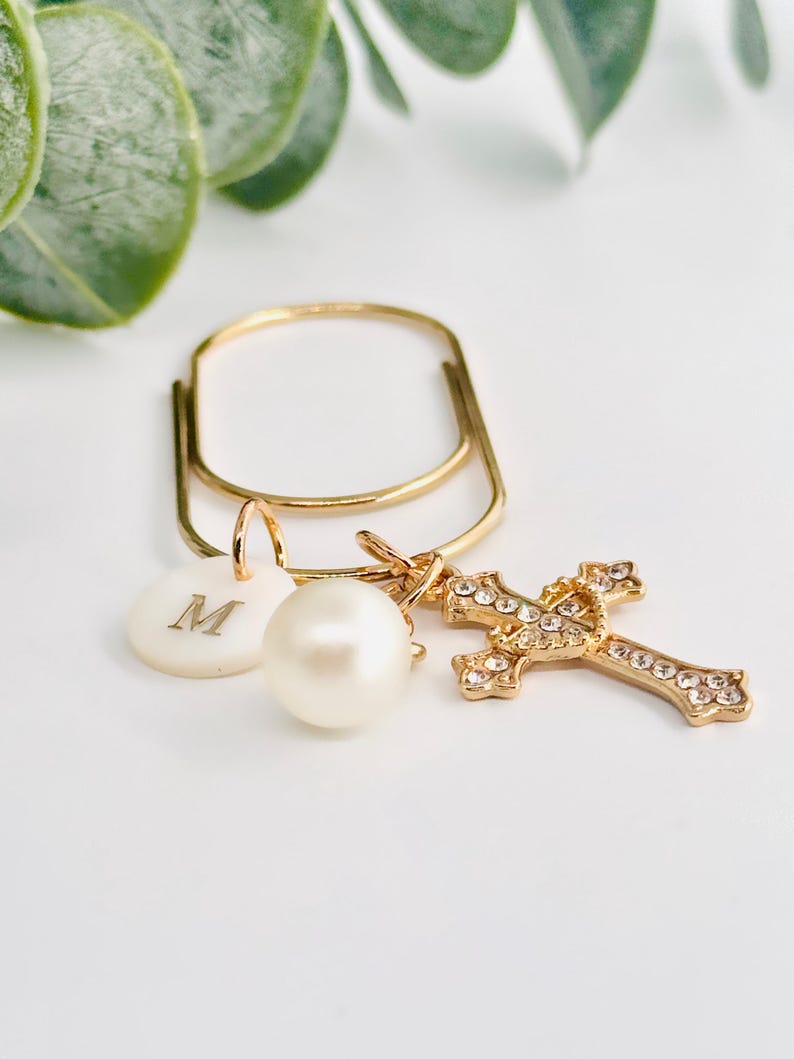 May include: Gold-toned keychain with a cross charm, a pearl, and a circular charm with the letter "M". The cross is encrusted with small, clear crystals. The keychain is set against a white background with green foliage.