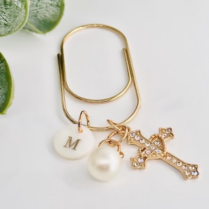 May include: A gold-colored paperclip with a white disc bearing the letter "M", a pearl, and a gold cross adorned with clear crystals. The cross and disc are attached to the paperclip with small gold rings.