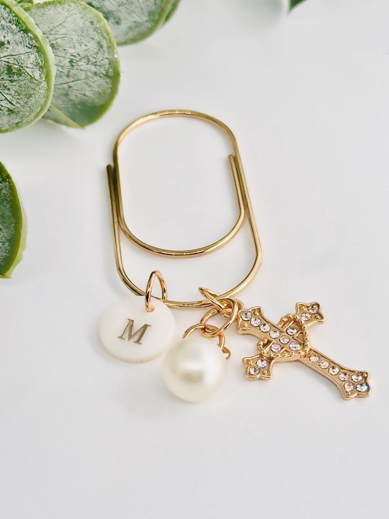May include: A gold-colored paperclip with a white disc bearing the letter "M", a pearl, and a gold cross adorned with clear crystals. The cross and disc are attached to the paperclip with small gold rings.