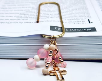 May include: Gold bookmark with a cross charm and pink and white beads. The bookmark is on top of a closed book with the text "Matthew 1:22" visible.