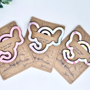 May include: Three wooden elephant-shaped bookmark clips with a pink, white and blue tie-dye design. Each clip has the text "I'll never forget your spot!" printed on it.