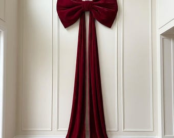 May include: A large, deep red velvet bow with two long, flowing tails that extend downwards. The bow features a central knot and wide, structured loops, creating a prominent decorative element mounted against a plain, light-coloured wall.