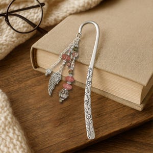 May include: A silver-toned metal hook bookmark featuring an engraved floral vine pattern along the stem. Attached to the top is a decorative charm cluster with pink oval beads, small clear crystals, and a detailed silver-toned angel wing charm.