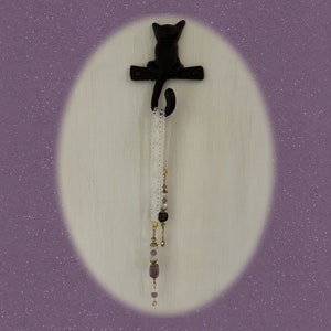 May include: A black metal cat hook with a white lace ribbon and purple and gold beads hanging from it. The hook is mounted on a white wall.