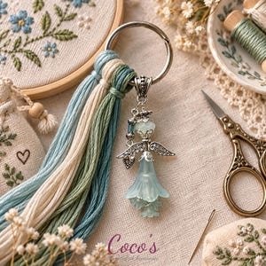 May include: A keychain featuring a silver-toned metal ring attached to a tassel made of light blue, cream, and sage green embroidery threads. A silver angel charm with a translucent pale blue bell-shaped skirt hangs from the metal ring.