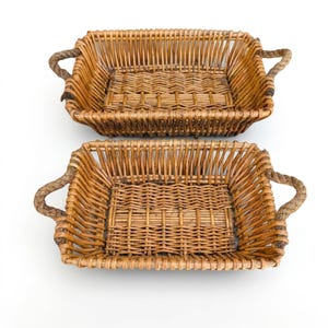 May include: Two rectangular woven wicker baskets with light brown natural finish and thick twisted rope handles on each side. The baskets feature an open weave pattern on the sides and a tighter, reinforced weave on the flat bottom base.