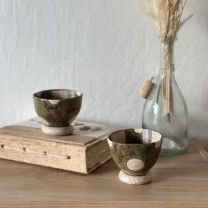 May include: Two small ceramic cups with a speckled green and brown glaze featuring circular cream-colored accents. Each cup has a distinct footed base with a speckled cream texture, showcasing a handcrafted appearance with organic shapes.