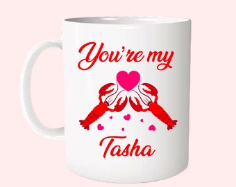 May include: White ceramic mug with a red and pink design. The mug features two red lobsters holding a pink heart with the text "You're my Tasha" printed on the mug.