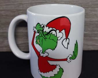 May include: White ceramic mug featuring a cartoon Grinch character in a Santa hat and red outfit with white trim. The Grinch is green with a grumpy expression. The mug has a white handle.