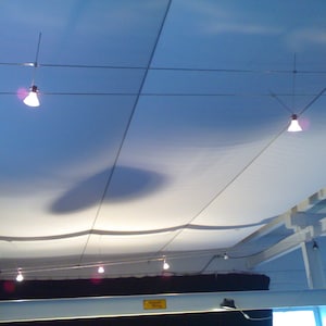 Può includere: Un soffitto in tessuto bianco con più apparecchi di illuminazione appesi a fili. Il tessuto è teso e sembra essere un elemento di design moderno.