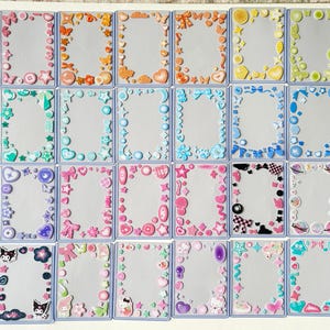 May include: A collection of thirty-two rectangular plastic card holders, each decorated with a unique border of colorful resin charms in various shapes including hearts, stars, bows, flowers, and moons, arranged in a grid on a white surface.