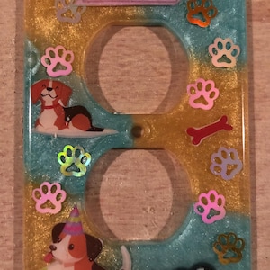 May include: A light blue and gold double outlet cover with a pink banner that says "dream big". The cover is decorated with paw prints, bones, and two cartoon dogs. One dog is wearing a party hat and holding a ball. The other dog is sleeping.
