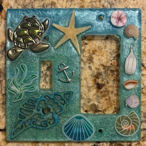 May include: A decorative light switch cover with a turquoise and teal resin base. It features a silver turtle, starfish, anchor, and various seashell designs. The cover has a glossy finish and is designed to fit a standard light switch.