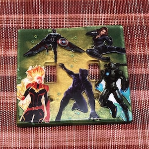 以下が含まれることがあります： 緑がかった薄い金色で、マーベルのスーパーヒーローのキャラクターのコラージュが施されたダブルライトスイッチカバー。キャラクターにはキャプテン・マーベル、ブラックパンサー、アイアンマン、ブラックウィドウが含まれています。カバーは星が散りばめられた金色の背景になっています。