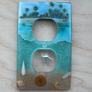 May include: Decorative electrical outlet cover with a tropical beach design. The cover depicts a blue and turquoise ocean, a sandy beach, seashells, and a small house. Palm trees and a boat are visible in the background.