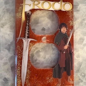 May include: A decorative electrical outlet cover featuring a design inspired by the Lord of the Rings. The cover is red with gold glitter accents and includes an image of Frodo, a sword, and the word "FRODO".