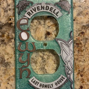 May include: A decorative teal-colored light switch cover with the word "Rivendell" at the top and "Last Homely House" at the bottom. The name "Eowyn" is vertically displayed in brown letters. The cover has a Celtic-inspired design on the right side.