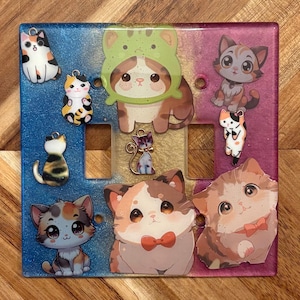 Peut inclure: Plaque décorative pour interrupteur avec des motifs de chats. La plaque est divisée en sections bleues, dorées et violettes. Plusieurs breloques et autocollants de chats sont attachés, présentant différentes races et styles de chats. Les chats sont dans diverses poses et couleurs.