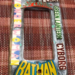 May include: A light switch cover with a silver background and colorful superhero logos. The logos include Superman, Wonder Woman, Flash, Green Lantern, and Cyborg. The bottom of the cover features a large Batman logo with a yellow background and blue accents.