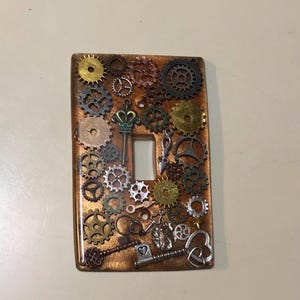 Może przedstawiać: Pokrywa włącznika światła z miedzianym tłem i różnymi metalowymi kołami zębatymi, kluczami i sercami. Pokrywa jest ozdobiona w stylu steampunk.