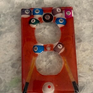 May include: A red, resin-coated light switch cover featuring a billiards theme. The cover has two openings and is decorated with pool balls numbered 6, 7, 8, 10, 12, 13, and the letter "L". Two pool cues are also visible.