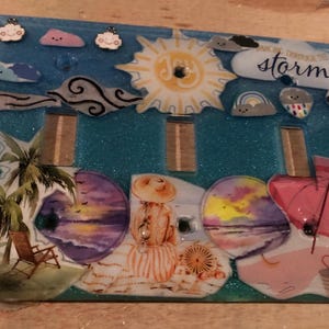 May include: A turquoise blue light switch cover with a beach theme. The cover features a sun, clouds, palm trees, an umbrella, and the text "We can weather the storm".