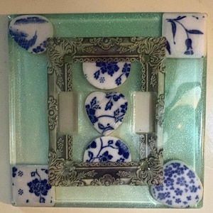 May include: Square glass coaster with a decorative silver frame. The coaster features blue and white floral patterned ceramic pieces, including a heart shape and semi-circles. The background is a light green color.