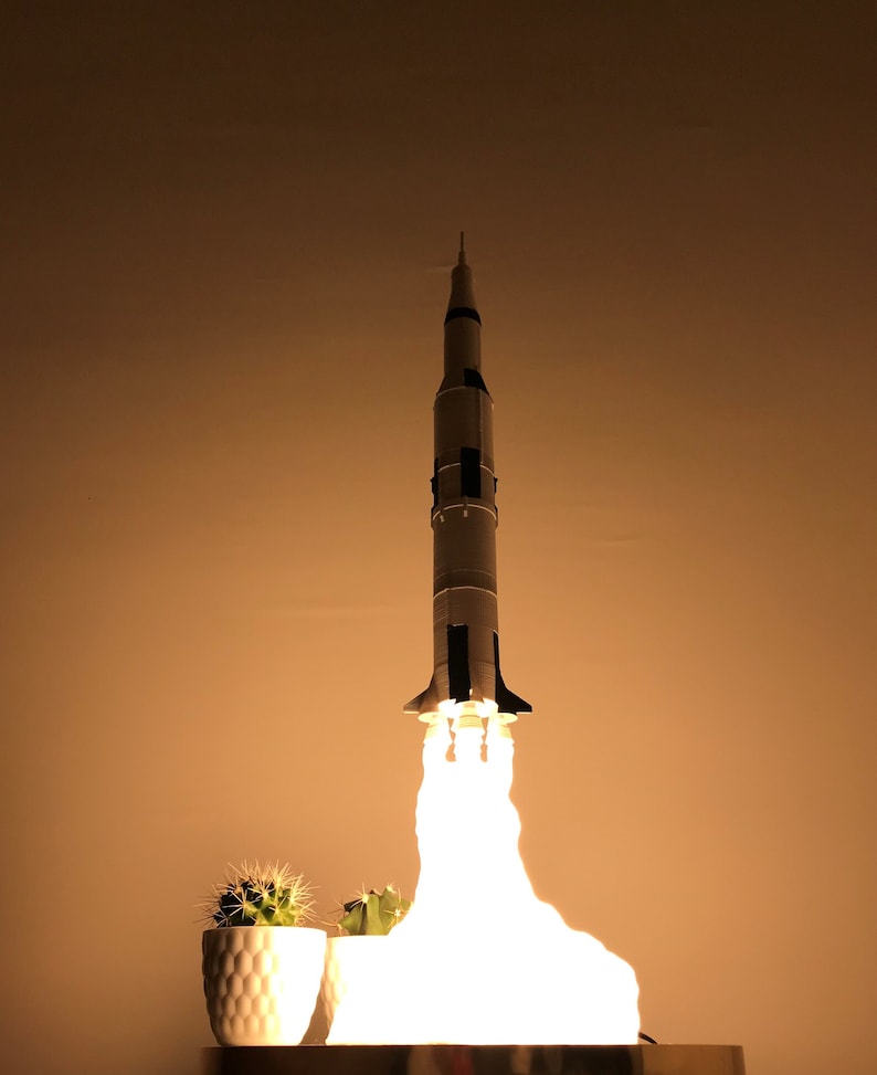 May include: A white model rocket with black accents is launching with a bright white light coming from the bottom. The rocket is on a table with a small potted cactus plant in front of it.