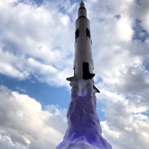 Pu&ograve; includere: Un razzo modello bianco e nero con una scia di fumo viola, che si lancia in un cielo blu con nuvole bianche.