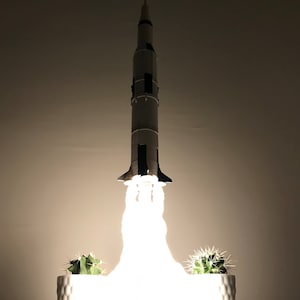 Pu&ograve; includere: Un modello di razzo bianco con accenti neri &egrave; illuminato, creando un bagliore bianco brillante. Il razzo &egrave; su un tavolo con due cactus in vaso davanti a lui.