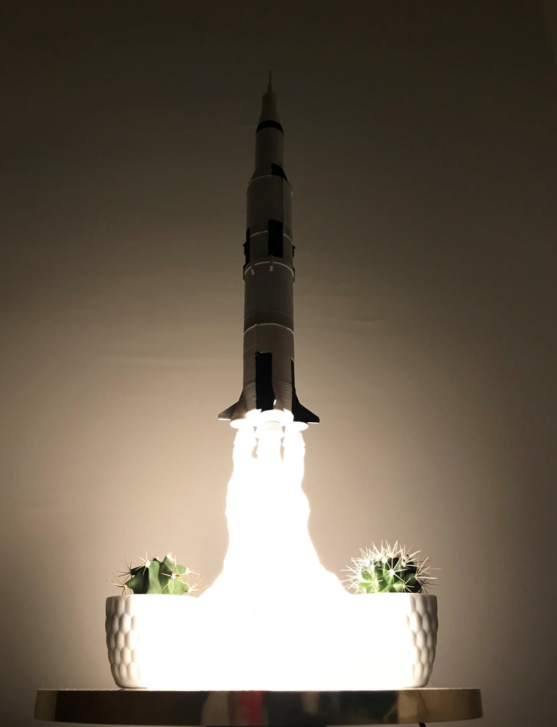 Pu&ograve; includere: Un modello di razzo bianco con accenti neri &egrave; illuminato, creando un bagliore bianco brillante. Il razzo &egrave; su un tavolo con due cactus in vaso davanti a lui.
