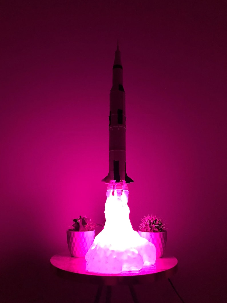 Pu&ograve; includere: Una lampada a forma di razzo bianco con un bagliore rosa, seduta su un tavolo con due piccole piante in vaso. Il razzo &egrave; illuminato dal basso, creando un bagliore rosa intorno a s&eacute;.