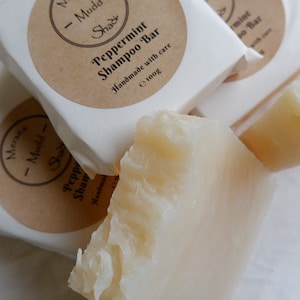 May include: Three white bars of peppermint shampoo soap, each wrapped in white paper with a brown label. The label reads "Meraca Mudd Shack" and "Peppermint Shampoo Bar" with the text "Handmade with care" and "e 100g".