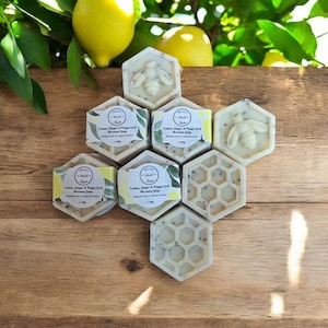 May include: Six hexagonal bars of handmade soap arranged in a honeycomb pattern. The soap is white with a light grey swirl and a bee design. The soap is labelled "Lemon, Ginger & Poppy Seed Bergamot Soap".