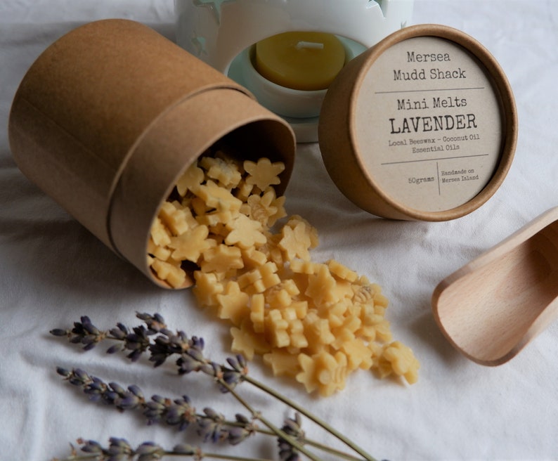 K&ouml;nnte beinhalten: Ein brauner Pappbeh&auml;lter mit Deckel, aus dem sternf&ouml;rmige Wachs-Schmelzen herausfallen. Die Wachs-Schmelzen sind hellgelb. Der Deckel hat ein Etikett mit der Aufschrift "Mersea Mudd Shack Mini Melts LAVENDER Local Beeswax - Coconut Oil Essential Oils Handmade on Mersea Island 50grams".