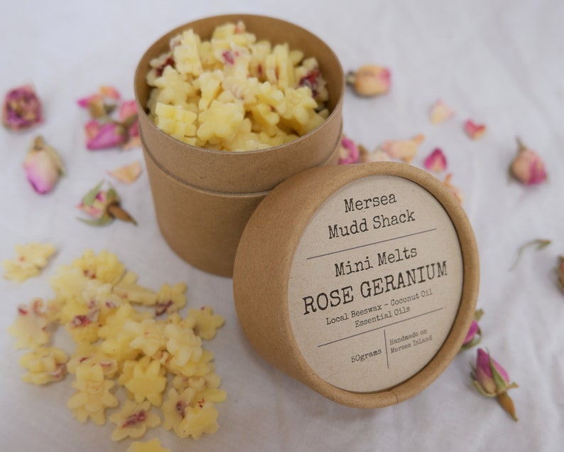 K&ouml;nnte beinhalten: Ein brauner Pappbeh&auml;lter mit einem wei&szlig;en Etikett, auf dem "Mersea Mudd Shack Mini Melts ROSE GERANIUM Local Beeswax - Coconut Oil Essential Oils Handmade on Mersea Island 50grams" steht. Der Beh&auml;lter ist mit kleinen, gelben Wachs-Schmelzen gef&uuml;llt, die wie Sterne geformt sind. Der Beh&auml;lter ist von getrockneten Rosenbl&auml;ttern umgeben.