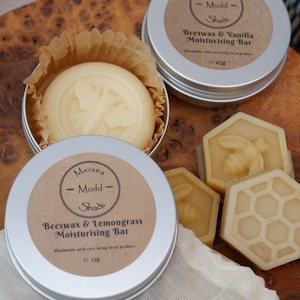 May include: Two round tins of beeswax and lemongrass and beeswax and vanilla moisturising bars. The tins are silver with brown labels. The labels have the text "Mersea Mudd Sha" and the product name. There are three hexagonal beeswax bars with a honeycomb design and a bee design.
